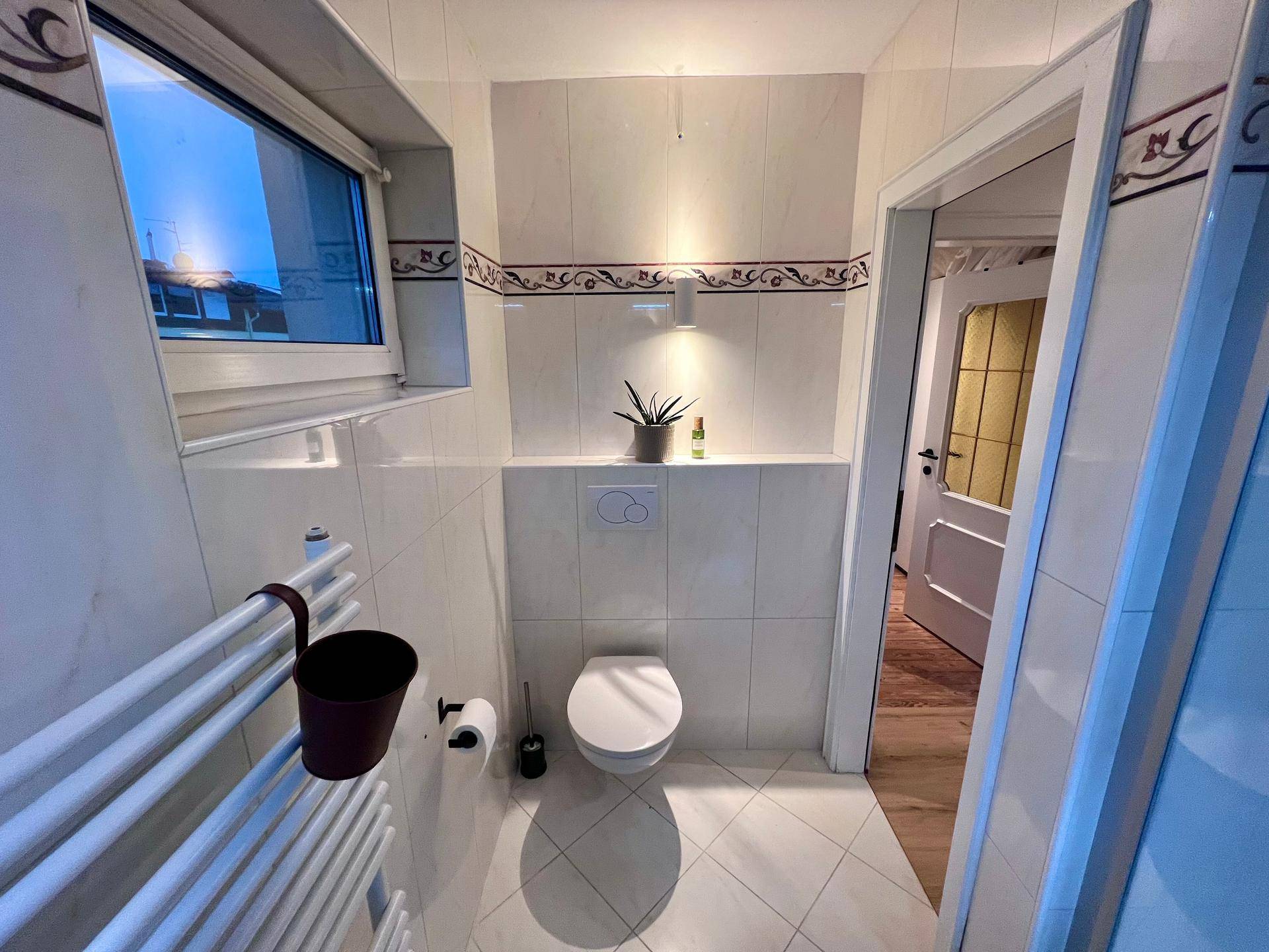 Gäste-WC mit Wand-WC, Fenster und Fliesenboden in hellem Design.