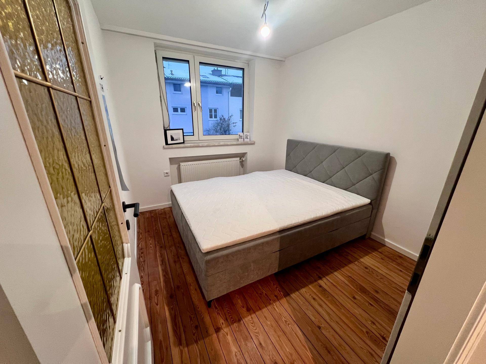 Helles Schlafzimmer mit Doppelbett, Holzboden und Fenster mit Blick nach draußen.