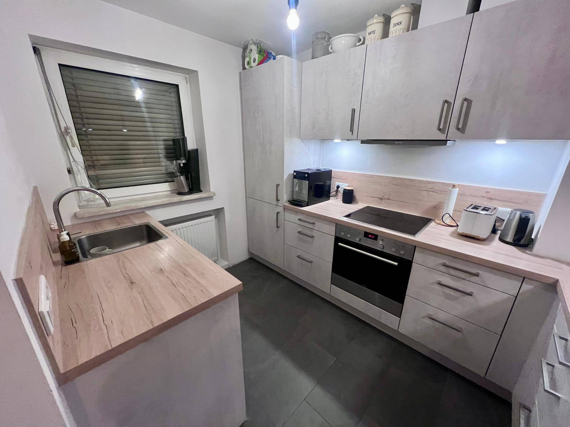 Funktionale Küche mit Spüle, Induktionskochfeld und Fenster mit Jalousien für natürliches Licht.