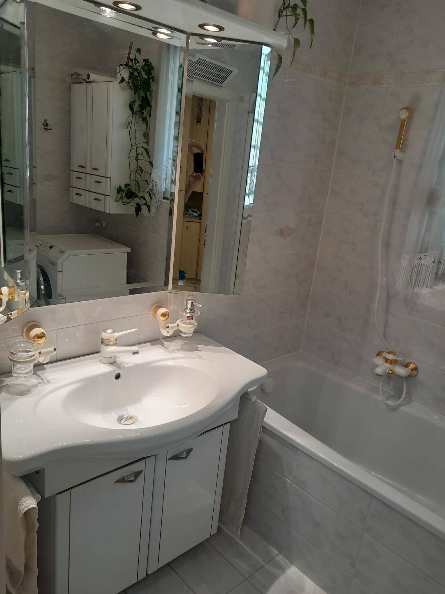 Badezimmer mit Waschbecken, Spiegelschrank, Badewanne und Waschmaschine, alles in hellen Fliesen gehalten.