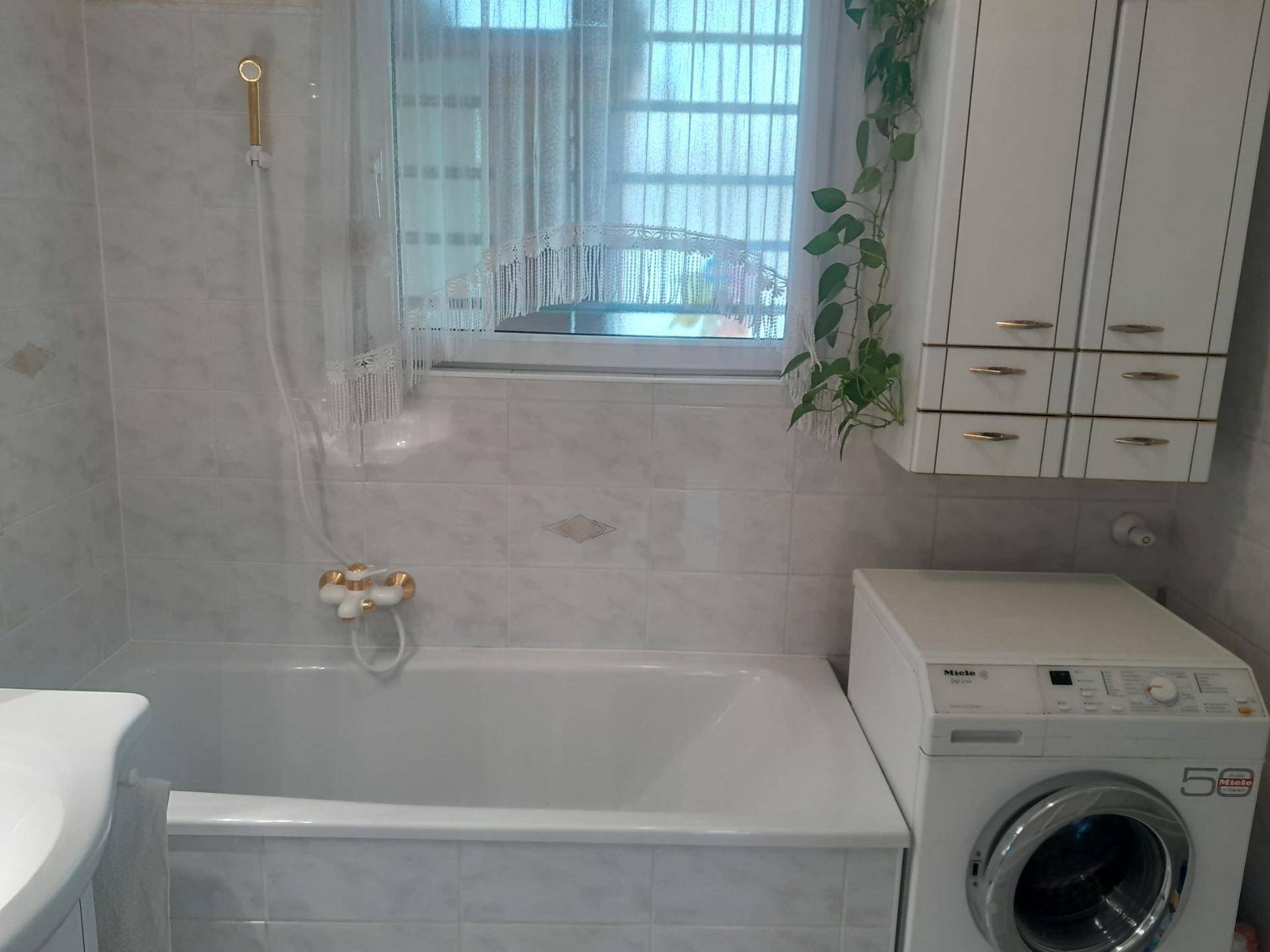 Badezimmer mit Badewanne, Waschmaschine, Fenster und einem praktischen Hängeschrank.
