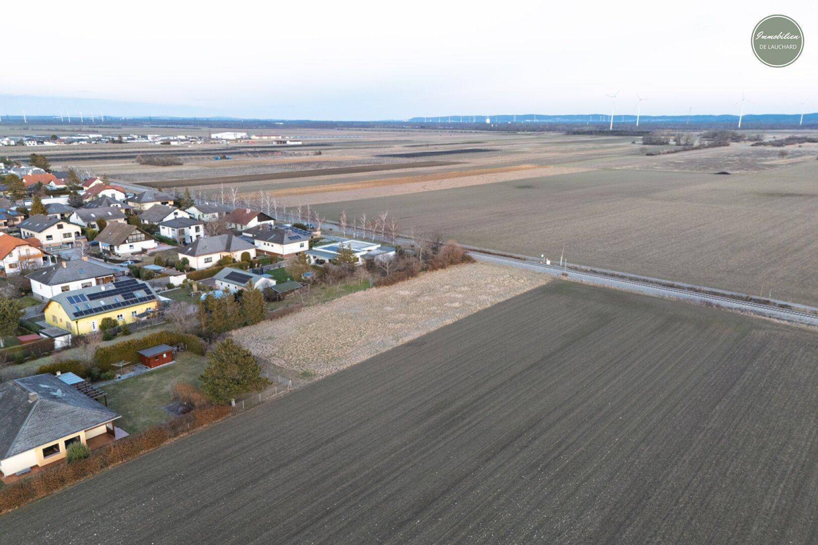 Weitläufige Luftaufnahme eines Grundstücks mit angrenzender Wohnbebauung und Windrädern am Horizont.