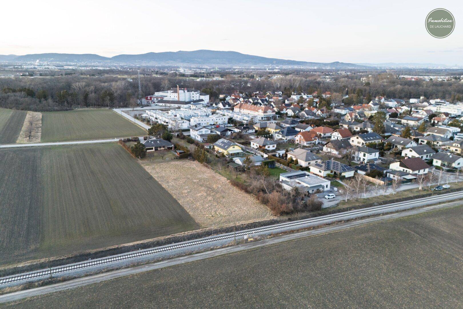 Luftaufnahme eines unbebauten Grundstücks neben Bahngleisen mit Blick auf die Wohnsiedlung und Berge.
