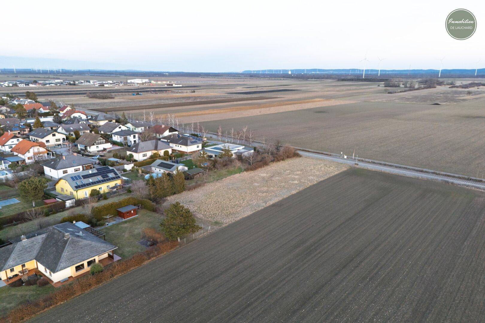 Weitläufige Luftaufnahme eines Grundstücks mit angrenzender Wohnbebauung und Windrädern am Horizont.