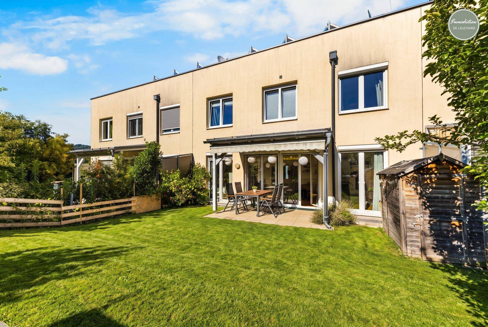 Hinterhof mit gepflegtem Rasen, Terrasse und Gartenhaus, umgeben von viel Grün.