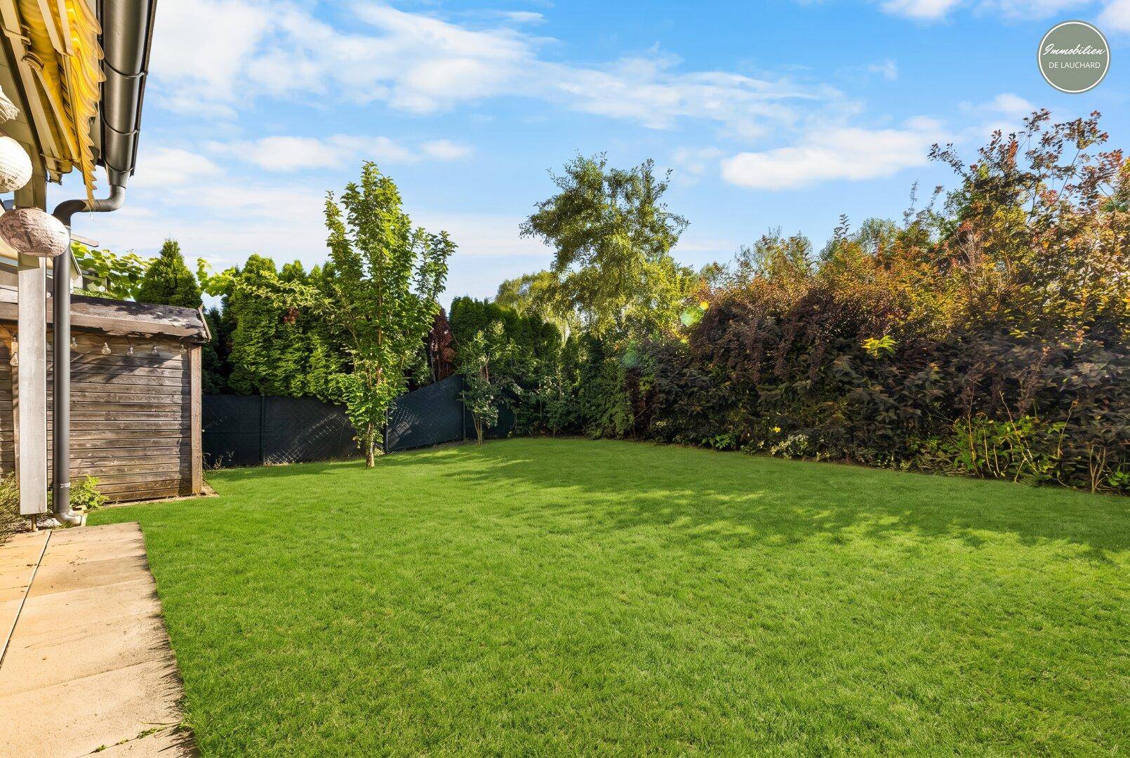 Gepflegter Garten mit Rasenfläche, Hecken und Gartenhaus unter blauem Himmel.