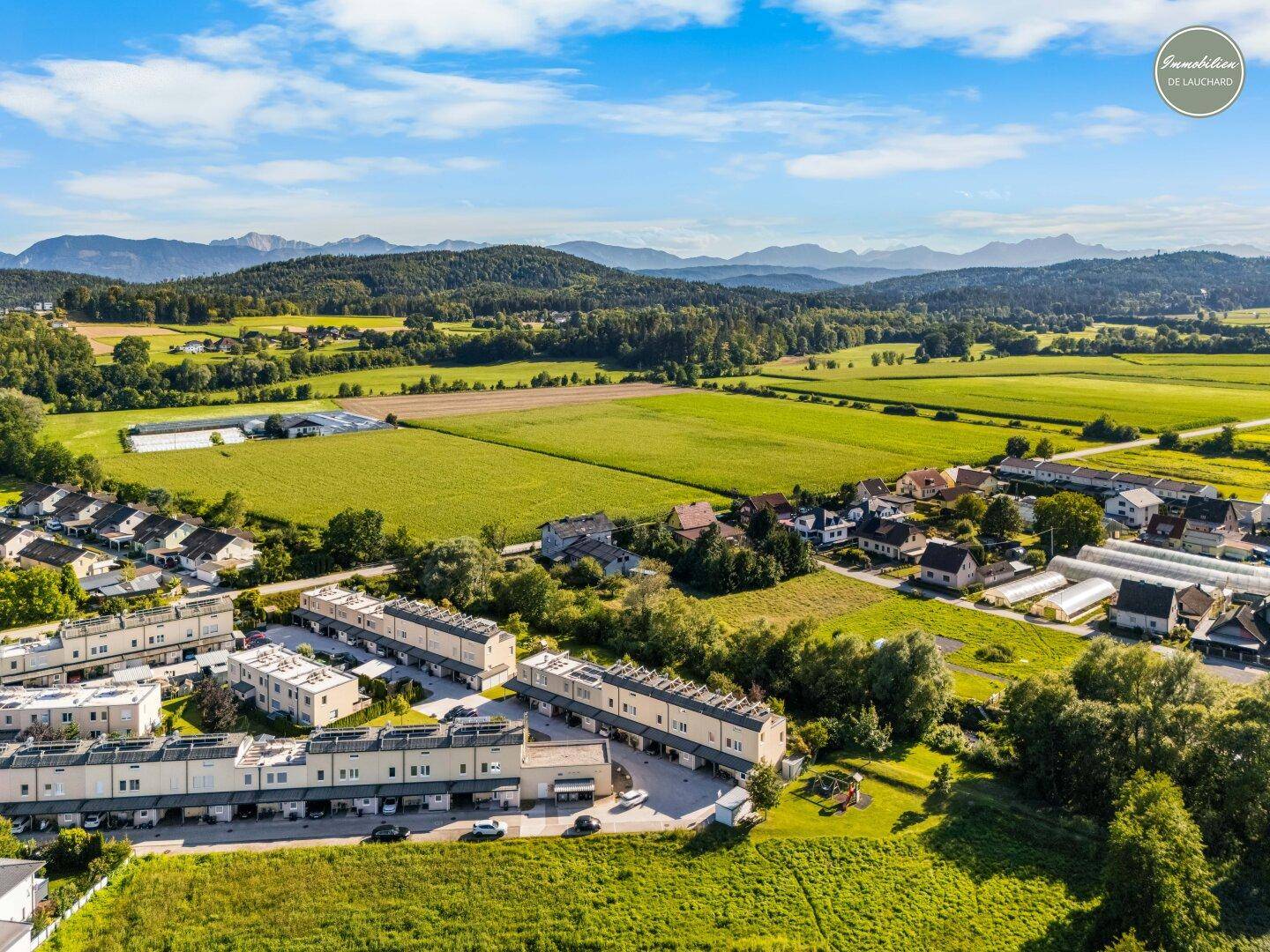 Weitläufige Luftaufnahme einer Wohnsiedlung eingebettet in grüne Felder mit Bergblick.