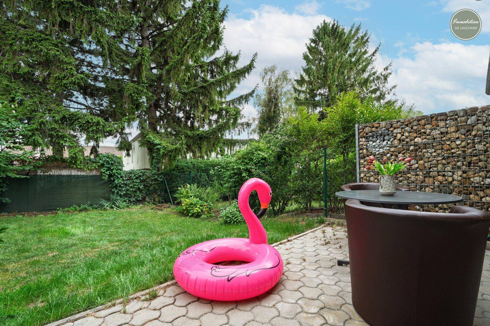 Grüner Gartenbereich mit gepflasterter Terrasse, Gabionenwand und hohen Bäumen.