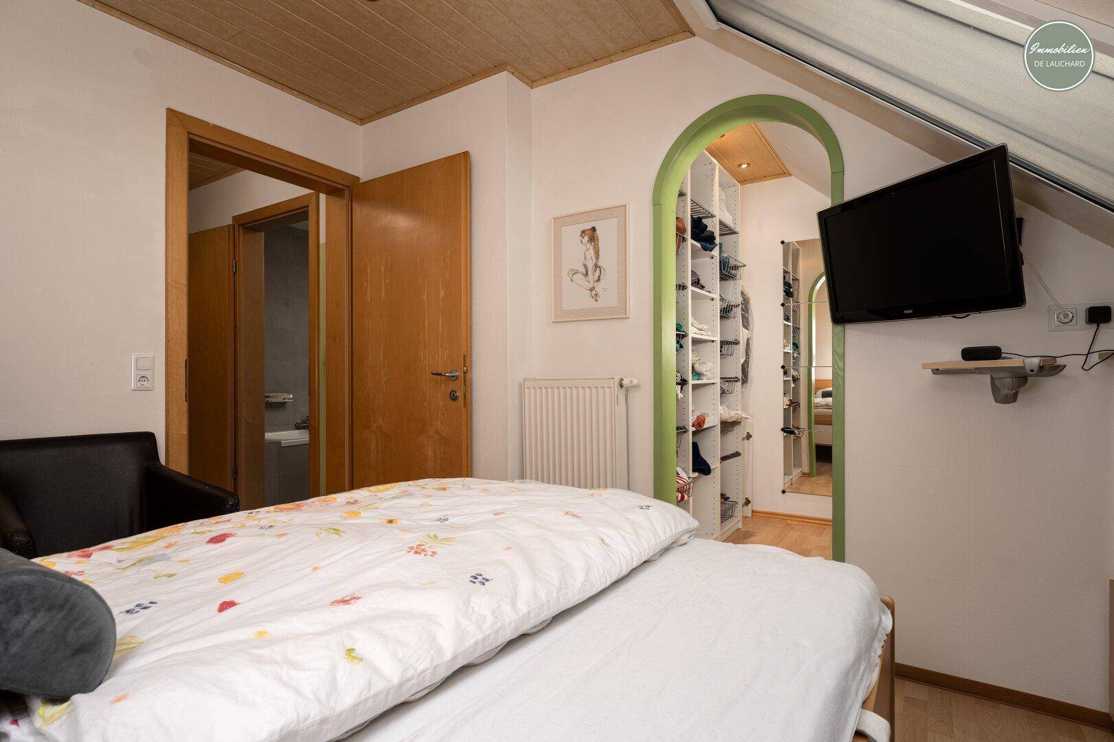 Helles Schlafzimmer mit Bett, Fernseher und Blick in den begehbaren Kleiderschrank.