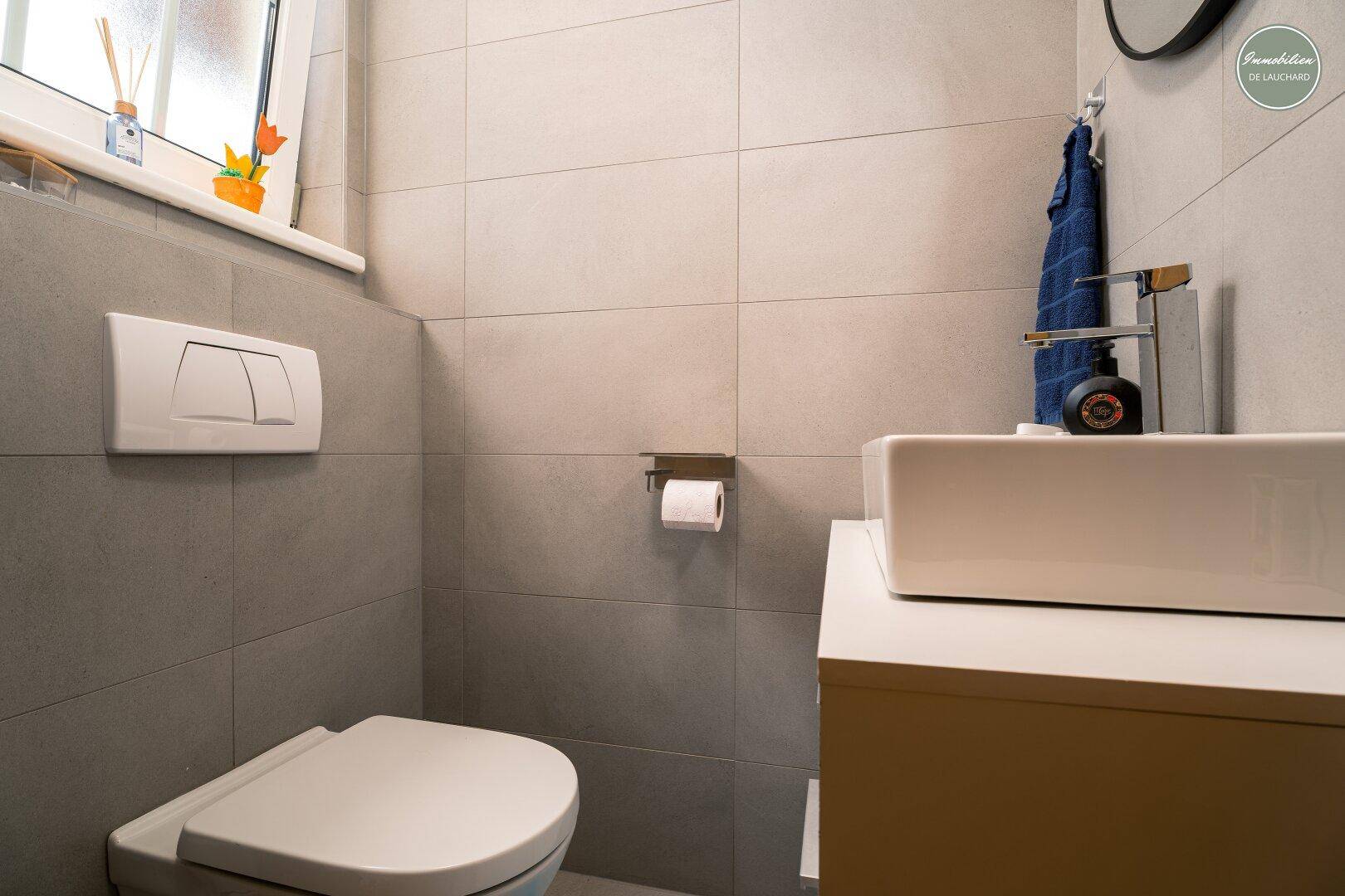 Modernes Gäste-WC mit grauen Fliesen, wandhängendem WC und Waschbecken unter einem Fenster.