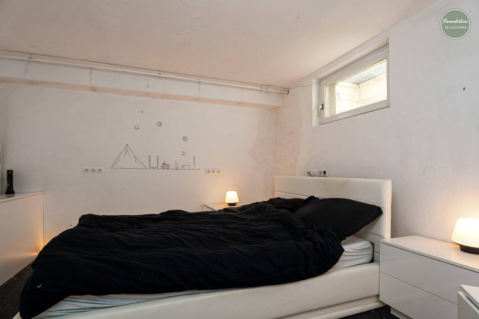 Schlafzimmer im Untergeschoss mit kleinem Fenster, weiß gestrichenen Wänden und dunkler Bettwäsche.