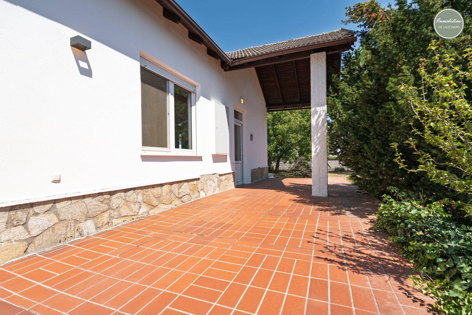 Gedeckte Terrasse mit Terrakottafliesen und Zugang zum Haus, umgeben von Grün.