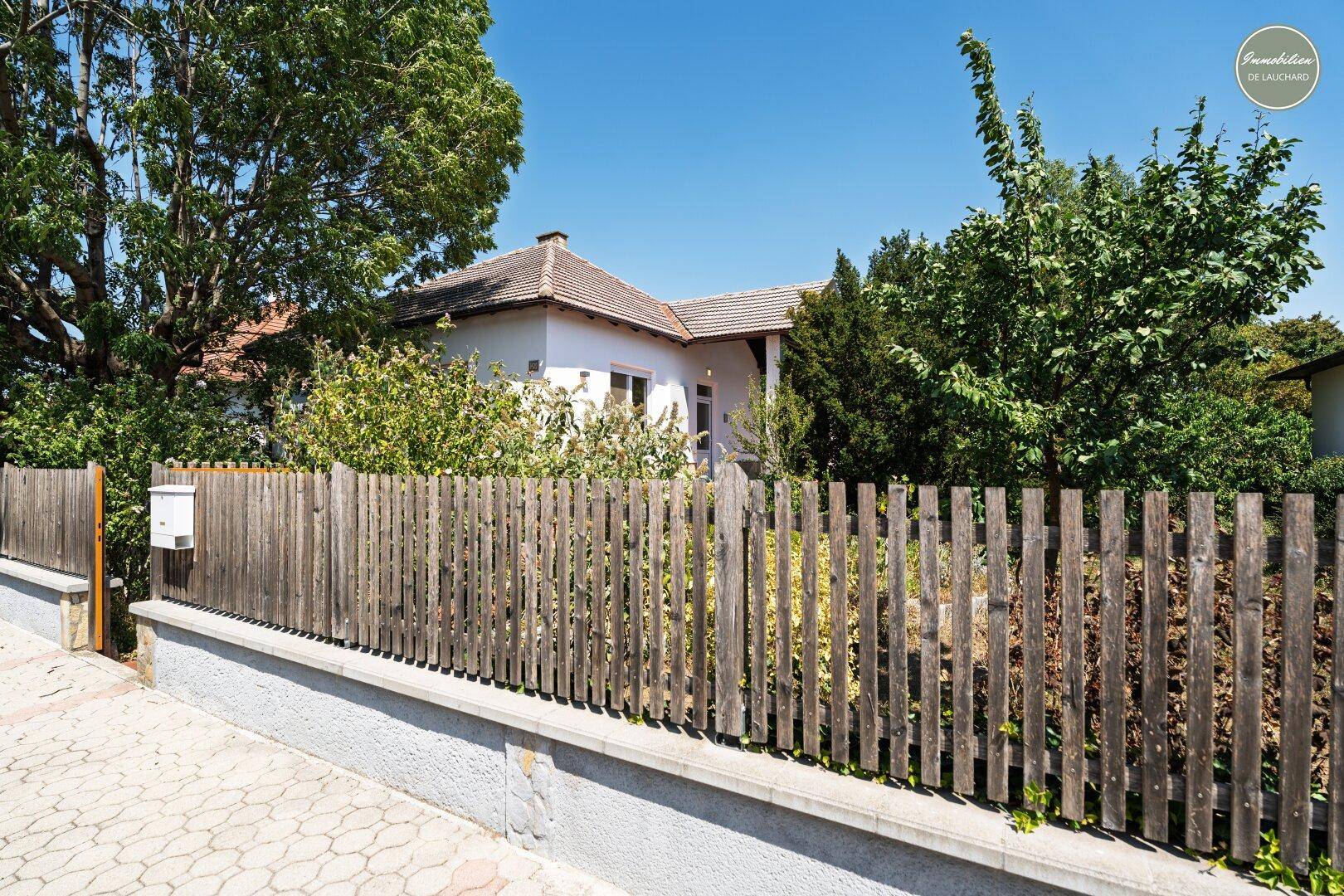 Einfamilienhaus von der Straße aus gesehen, hinter einem Holzzaun mit viel Grün und Bäumen.