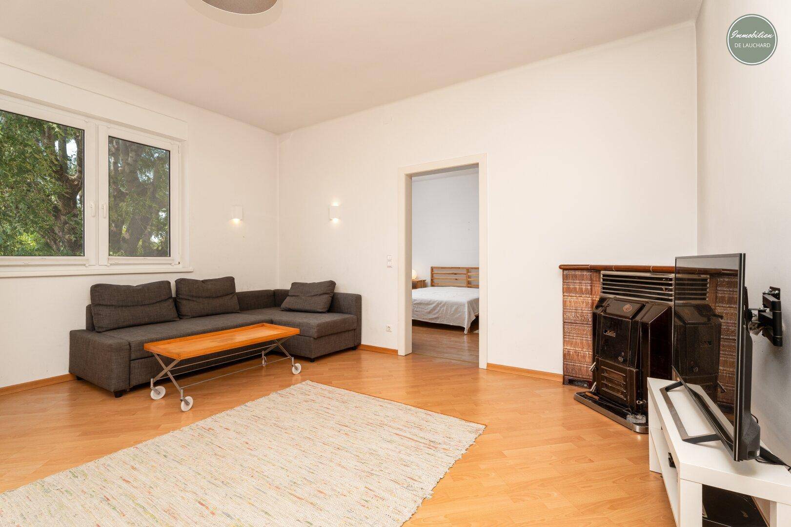 Gemütliches Wohnzimmer mit Sofa, Holzboden und einem Kaminofen, angrenzend an ein Schlafzimmer.