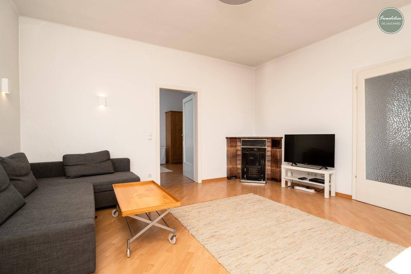 Wohnzimmer mit hellem Holzboden, bequemer Couch und Blick auf den Kaminofen und eine Tür.