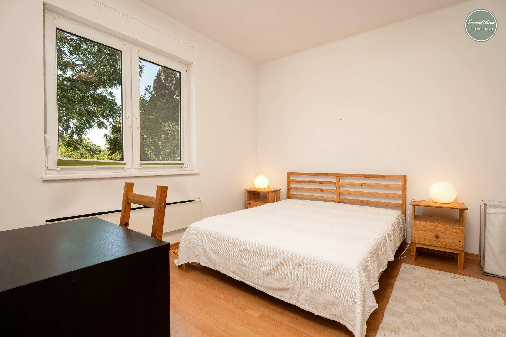Helles Schlafzimmer mit Doppelbett, Holzmöbeln und Blick ins Grüne durch das Fenster.