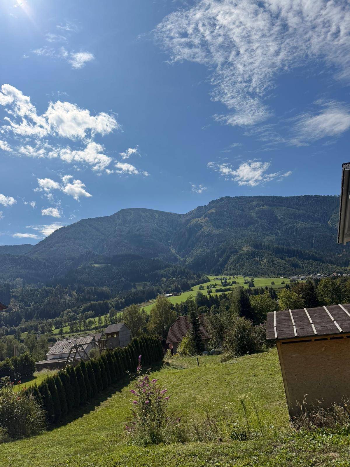 Weitläufiger Blick über eine grüne Berglandschaft mit vereinzelten Häusern unter blauem Himmel.