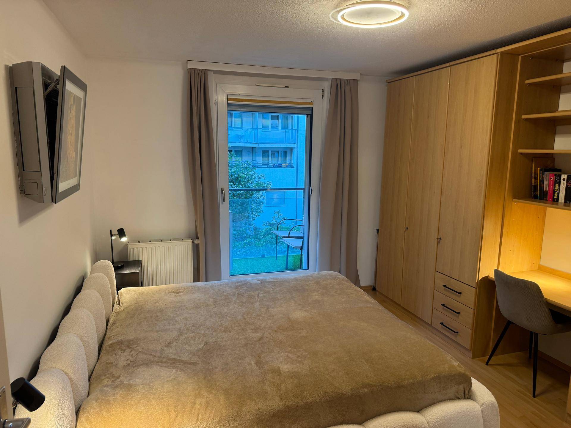 Schlafzimmer mit großem Bett, Holzschrank und Schreibtisch, mit Balkonzugang.