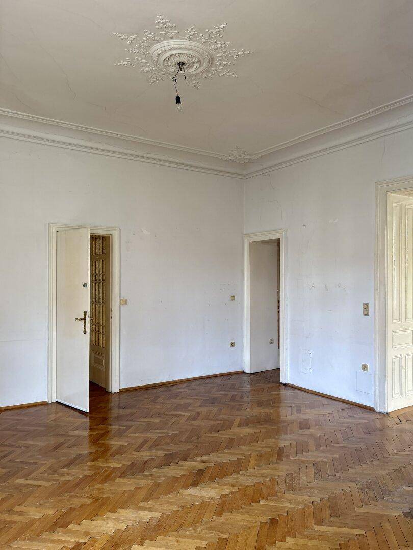 Geräumiges Wohnzimmer mit klassischem Fischgrätparkett und hohen Türen, das viel Potenzial bietet.