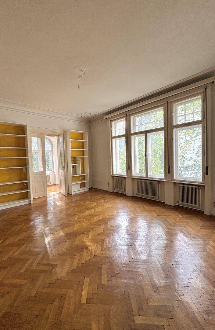 Heller Wohnraum mit großen Fenstern, Fischgrätparkett und eingebauten Regalen, die viel Stauraum bieten.