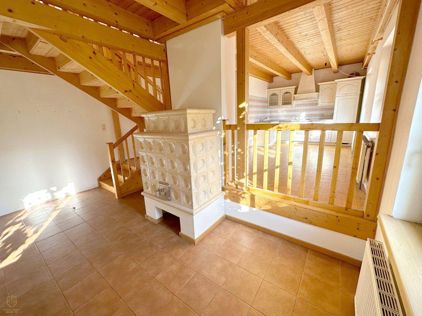 Heller Wohnbereich mit traditionellem Kachelofen, Holztreppe und sichtbaren Deckenbalken.