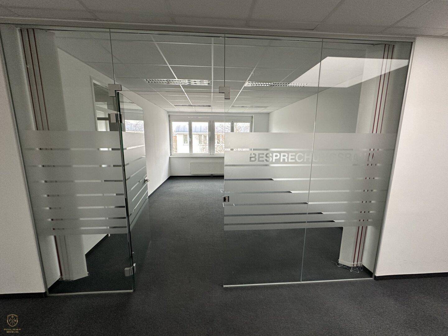 Blick durch eine satinierte Glastrennwand in einen hellen Büroraum mit Teppichboden und Fenstern.