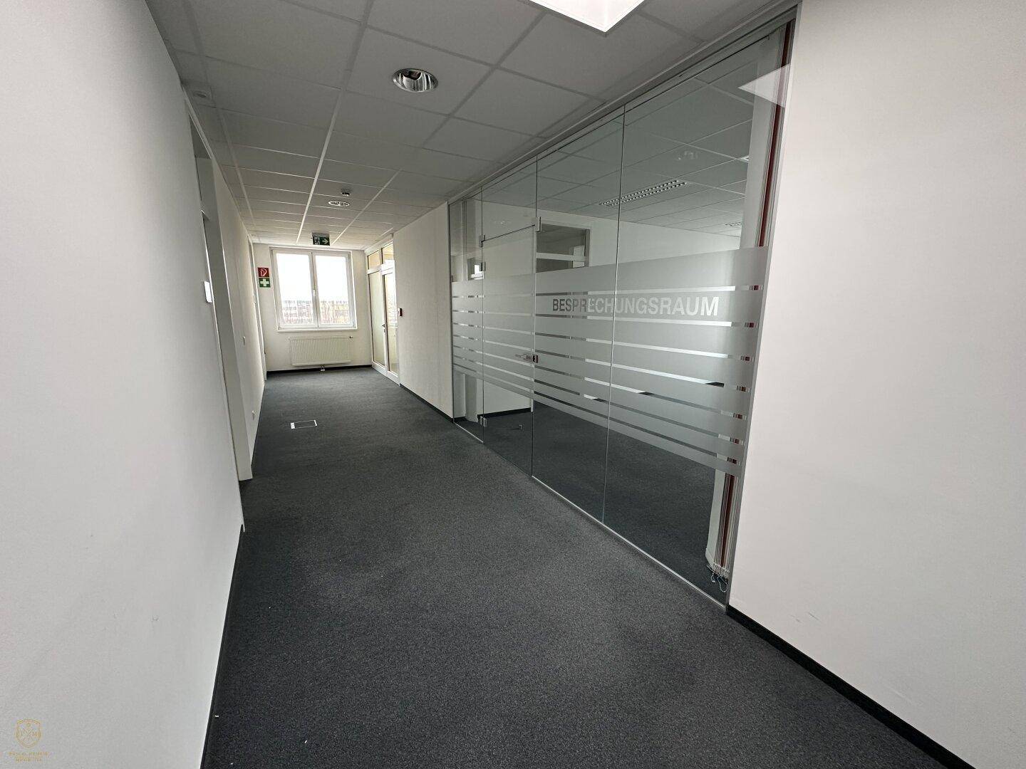 Büroflur mit Teppichboden und einer modernen Glastrennwand, die zu einem Besprechungsraum führt.