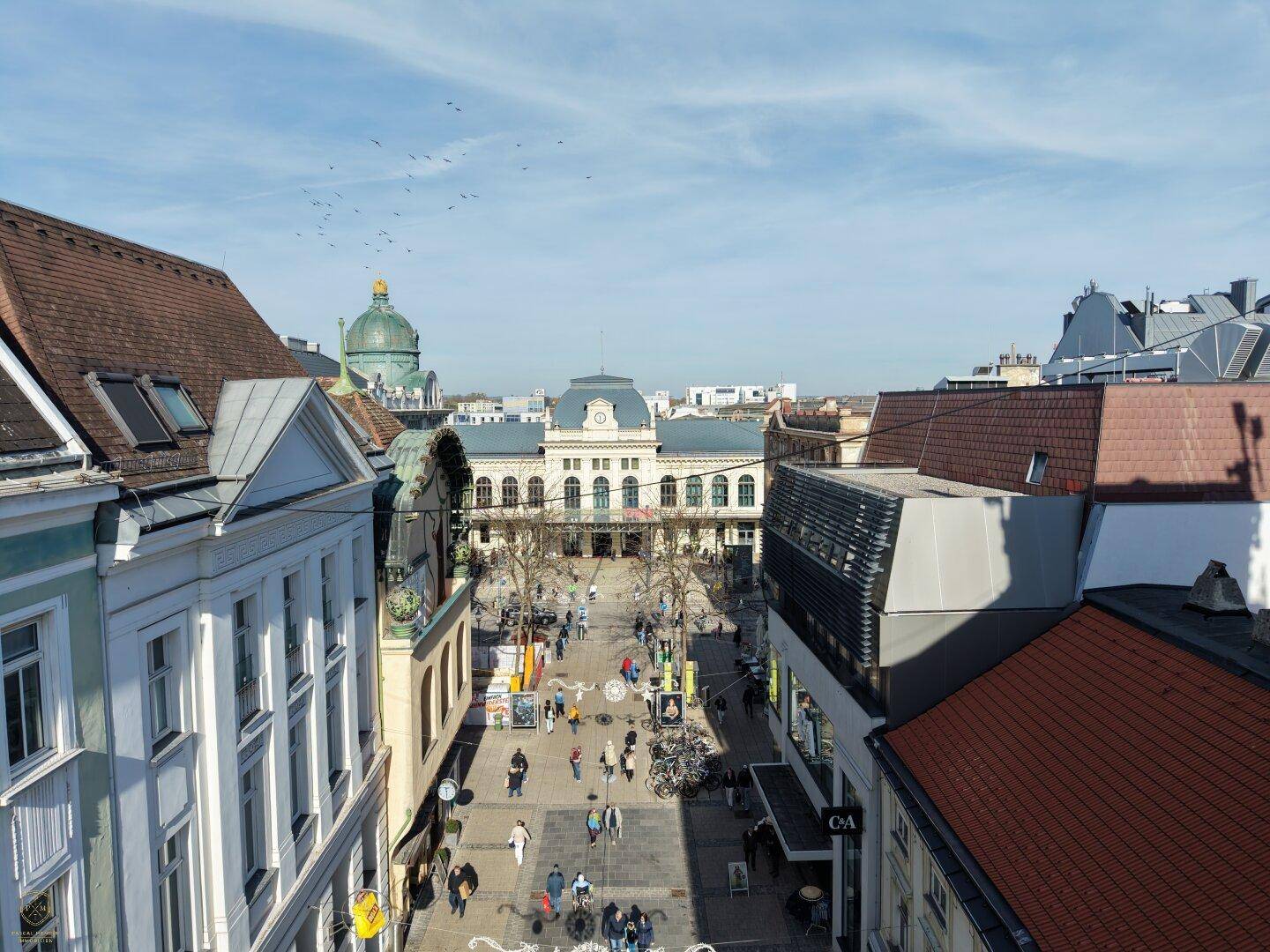 Panoramablick über eine belebte Stadtstraße mit historischen Gebäuden und einem großen Platz im Hintergrund.