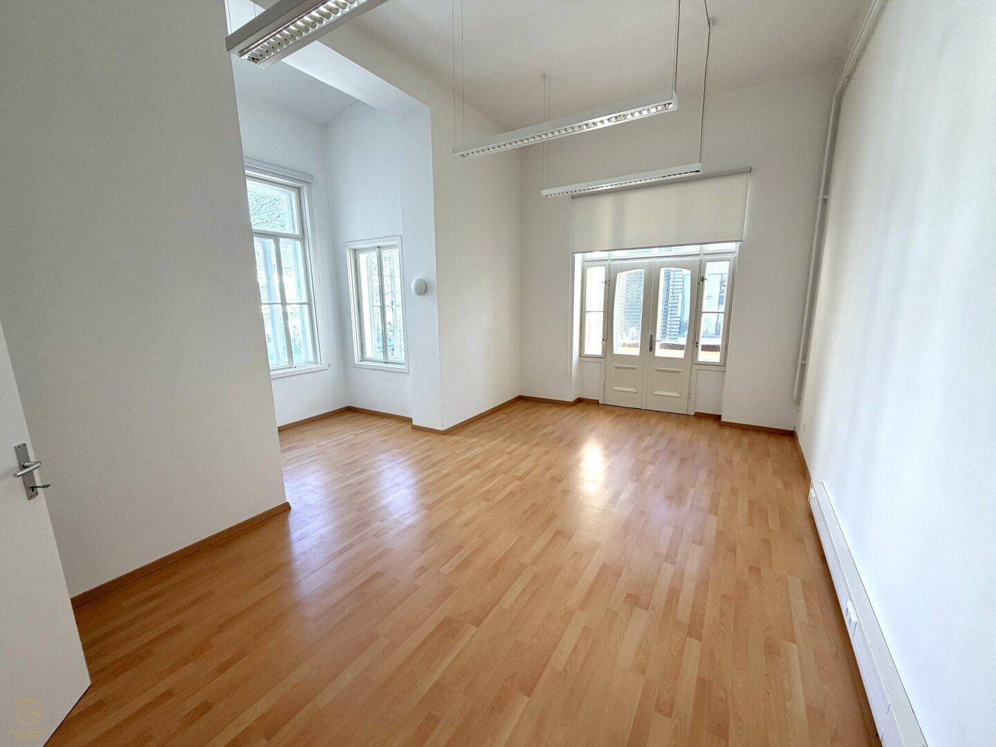 Großer, heller Raum mit Laminatboden, hohen Fenstern und modernen Deckenleuchten.
