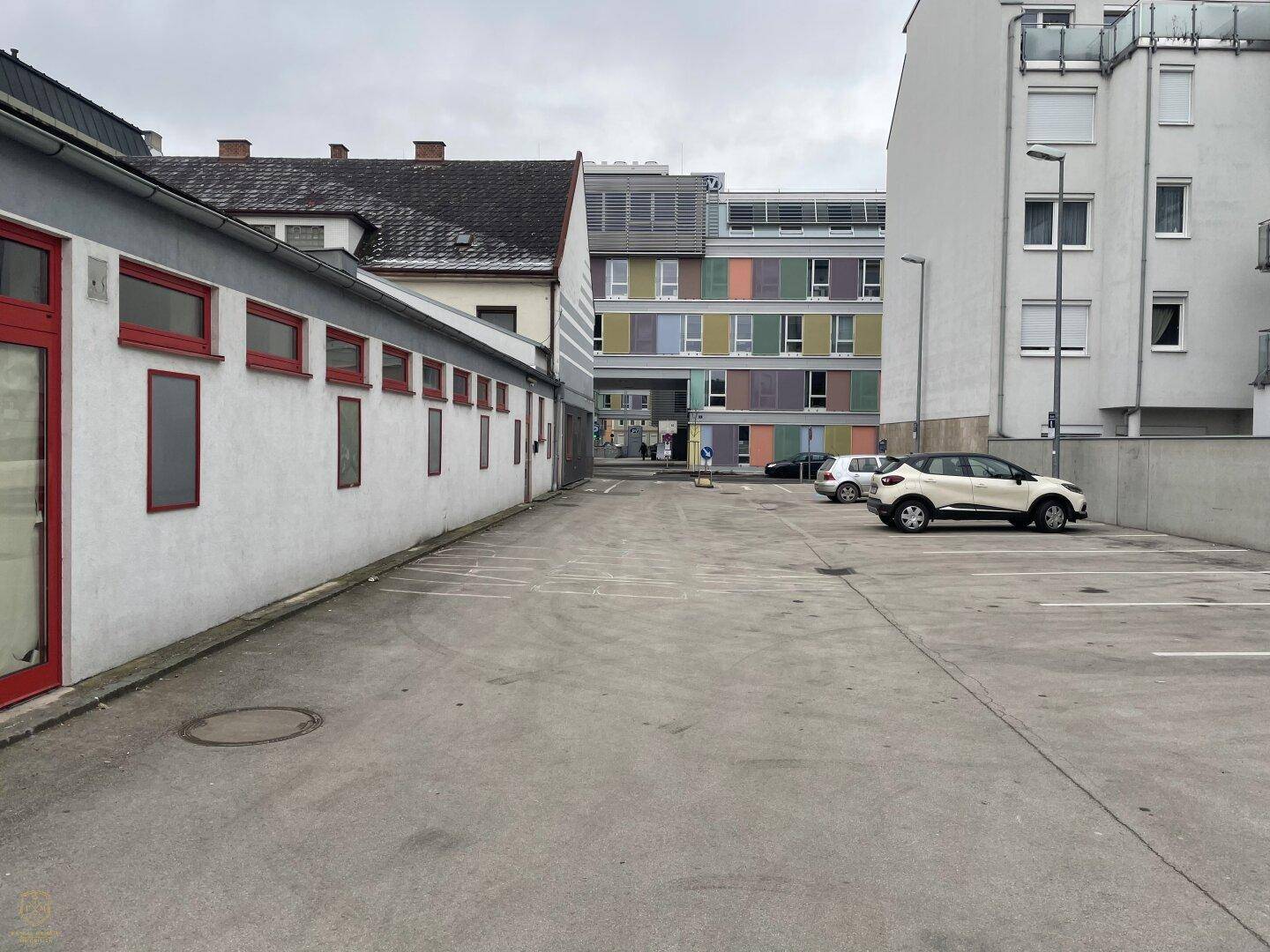 Großer Außenparkplatz neben dem Gebäude mit weiteren Gebäuden im Hintergrund und einem hellen Himmel.