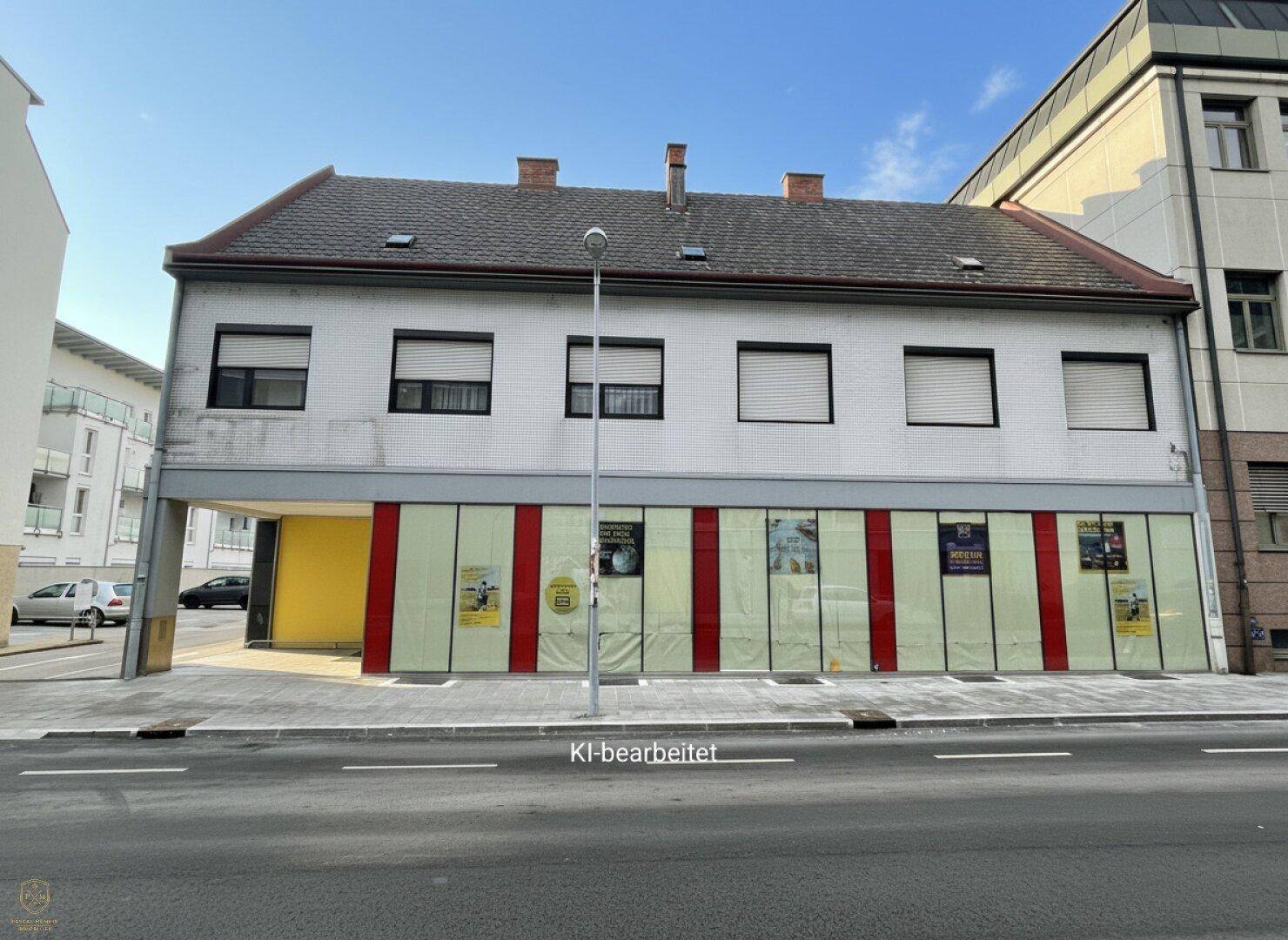 Straßenansicht des Gebäudes mit Ladenfronten im Erdgeschoss und geschlossenen Rollläden an den oberen Fenstern.
