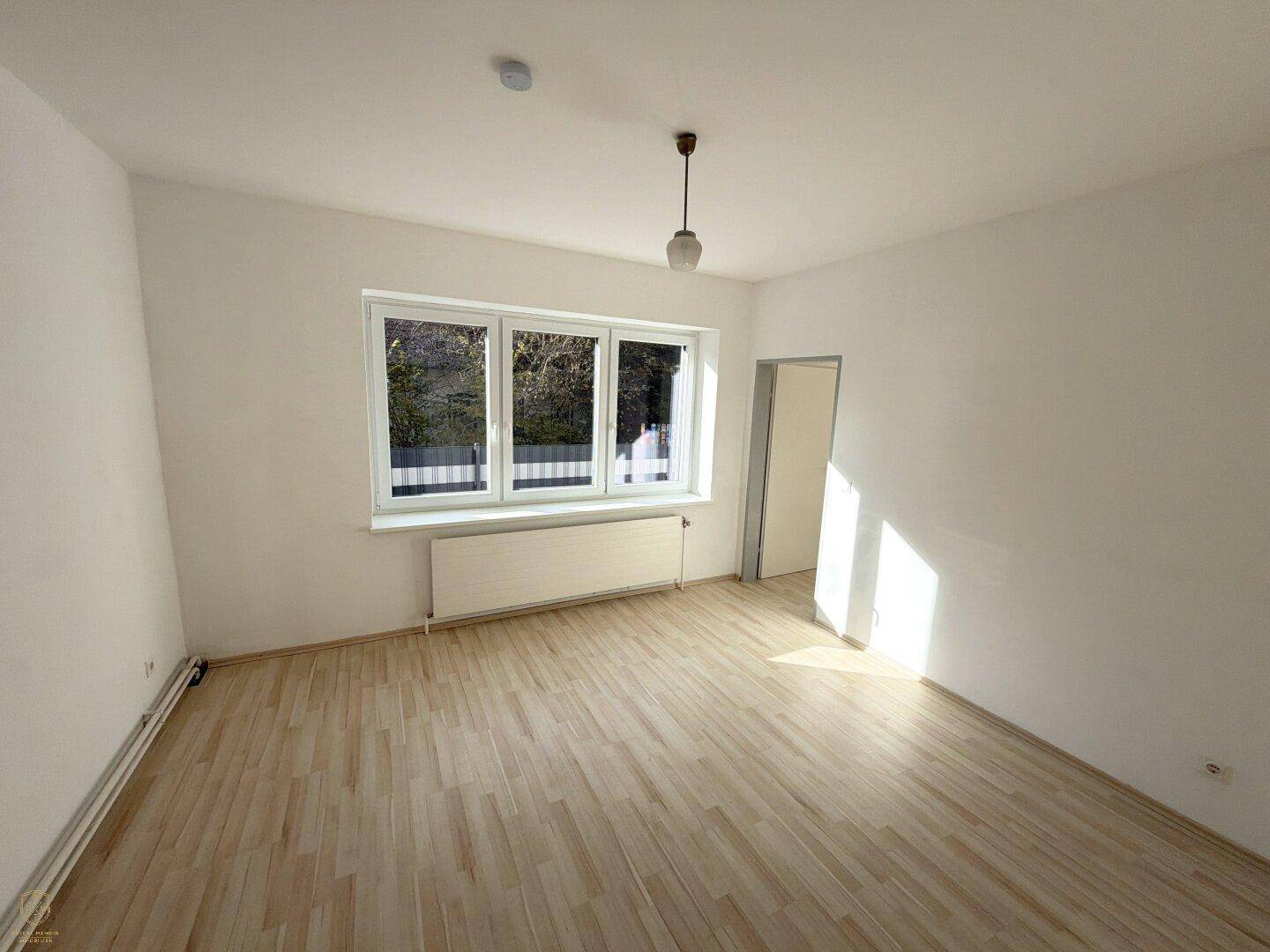 Lichtdurchfluteter Raum mit hellem Holzboden und großen Fenstern, die einen grünen Ausblick bieten.