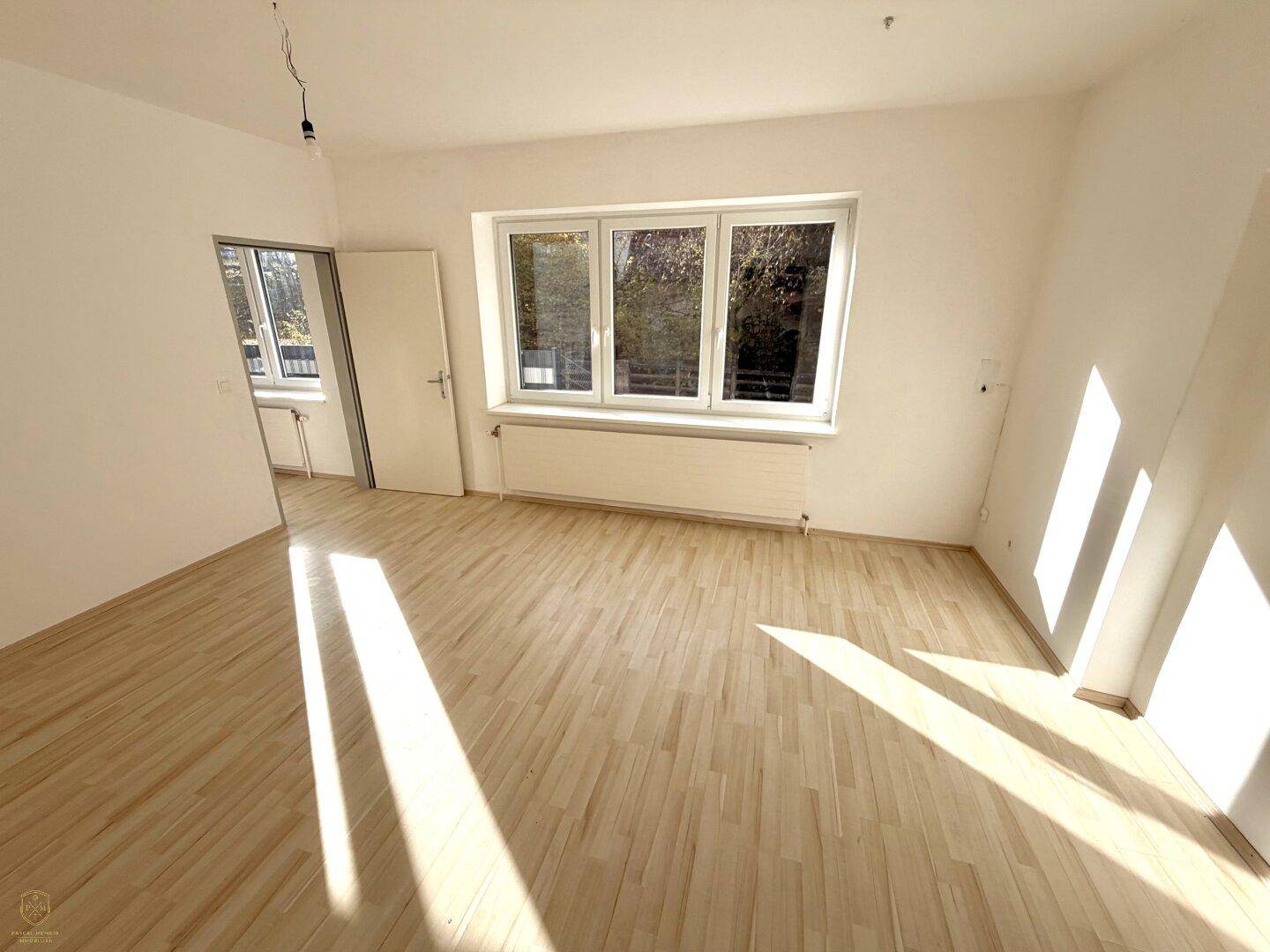 Heller Wohnraum mit Holzboden und großen Fenstern, die viel Sonnenlicht hereinlassen und den Raum erhellen.