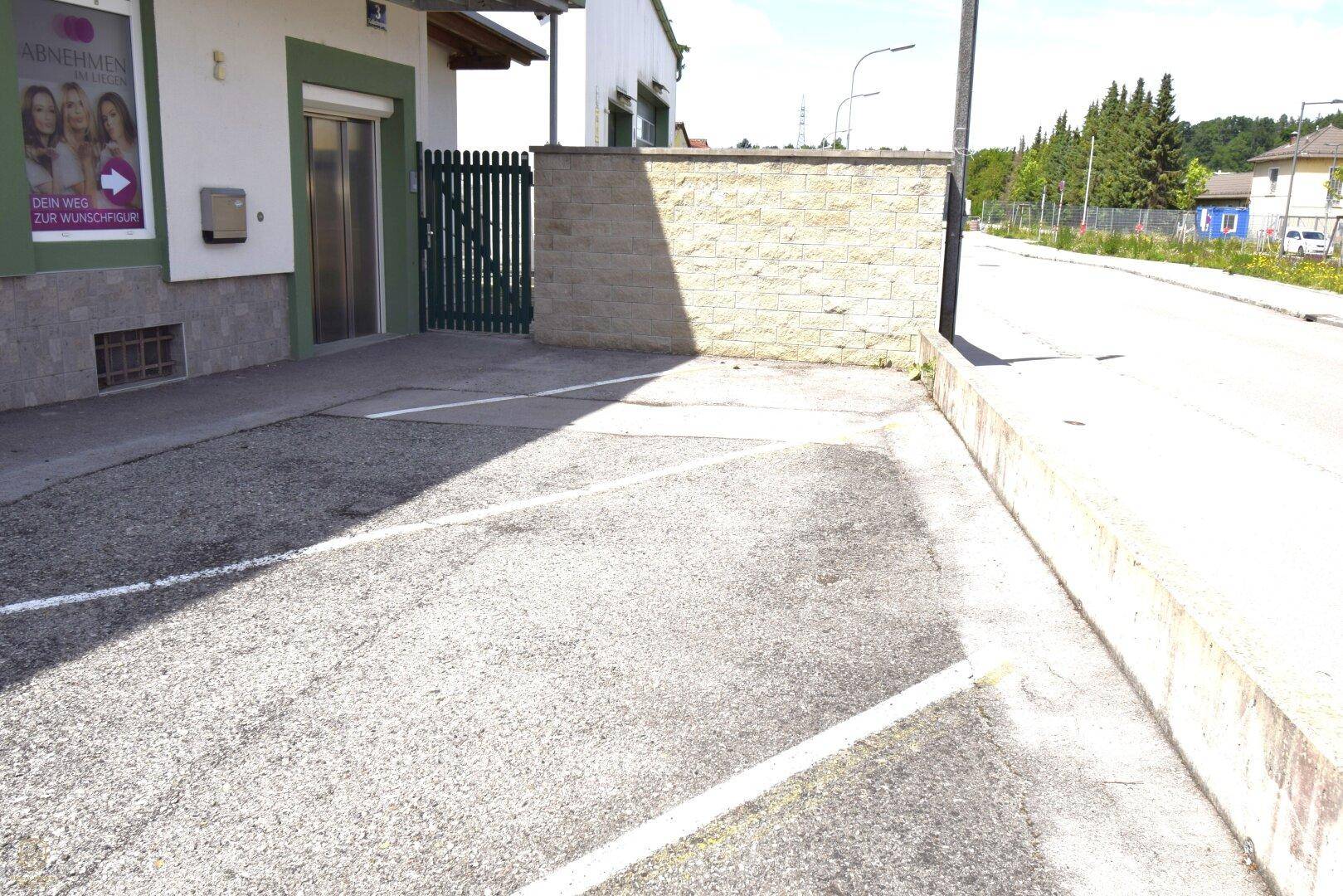 Außenparkplatz vor einem Gebäude mit Werbeschild und einer grünen Pforte.
