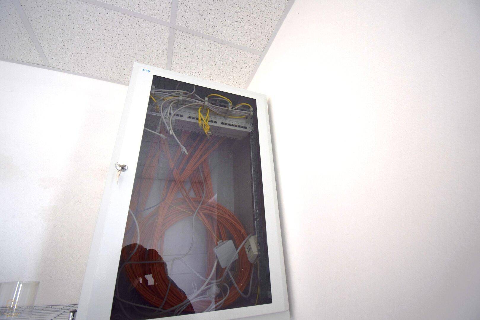 Technischer Raum mit einem Serverschrank voller Kabel und einer Rasterdecke.