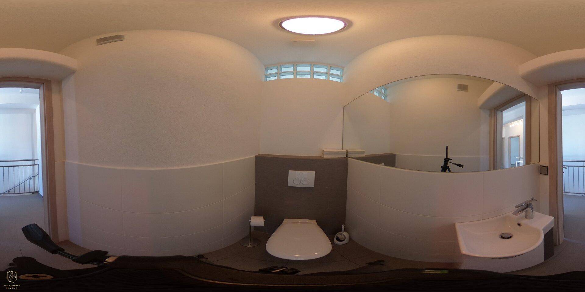 Modernes Badezimmer mit wandhängender Toilette, Waschbecken und einem runden Deckenlicht, das den Raum erhellt.