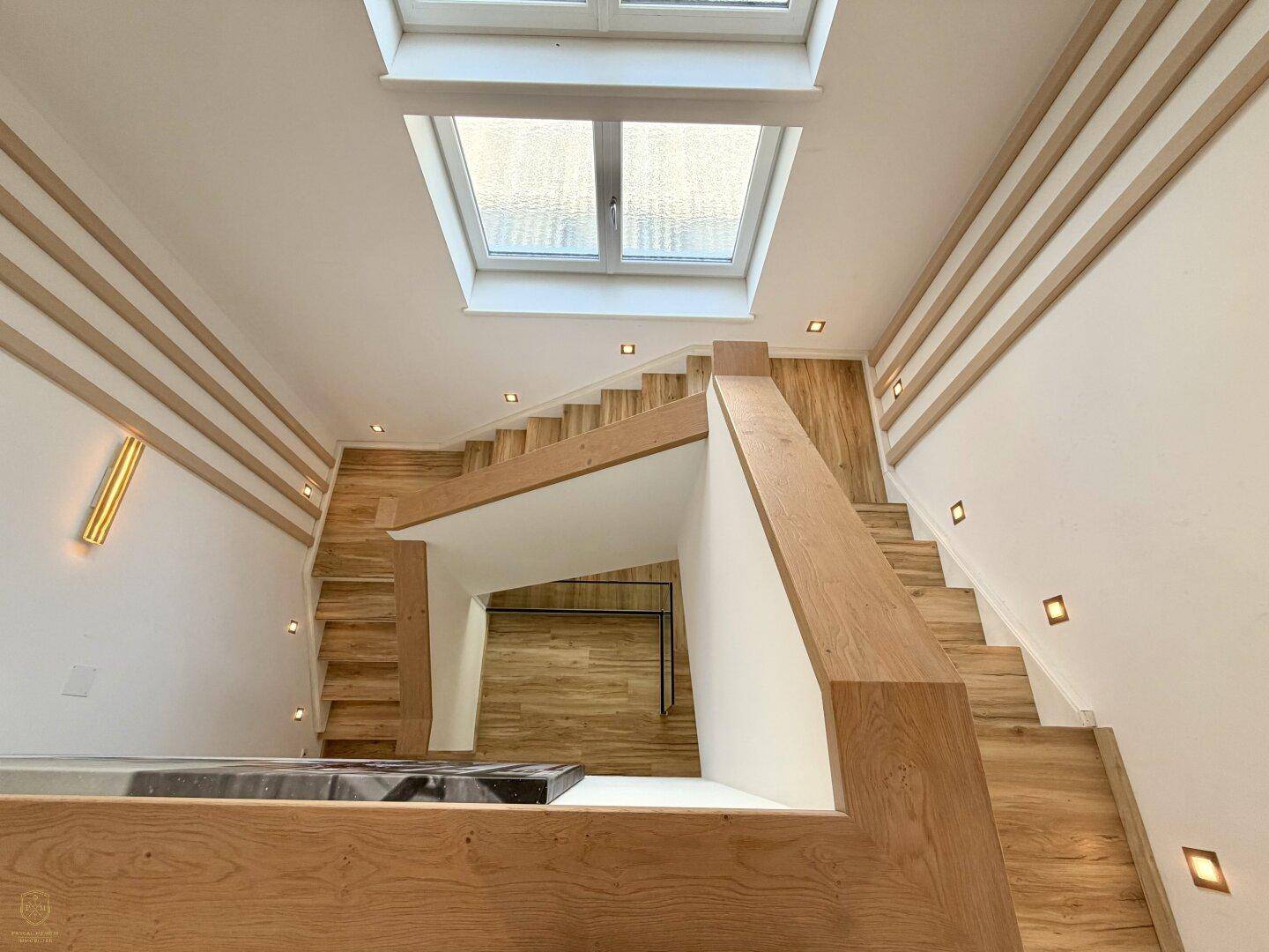 Helles Treppenhaus mit Holzstufen, modernen Geländern und einem großen Dachfenster.
