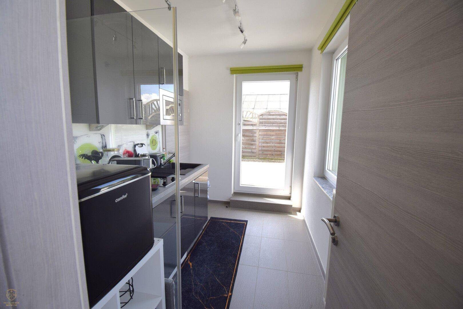 Schmale Küche mit grauen Hochglanzschränken, kleinem Kühlschrank und Zugang zum Außenbereich.
