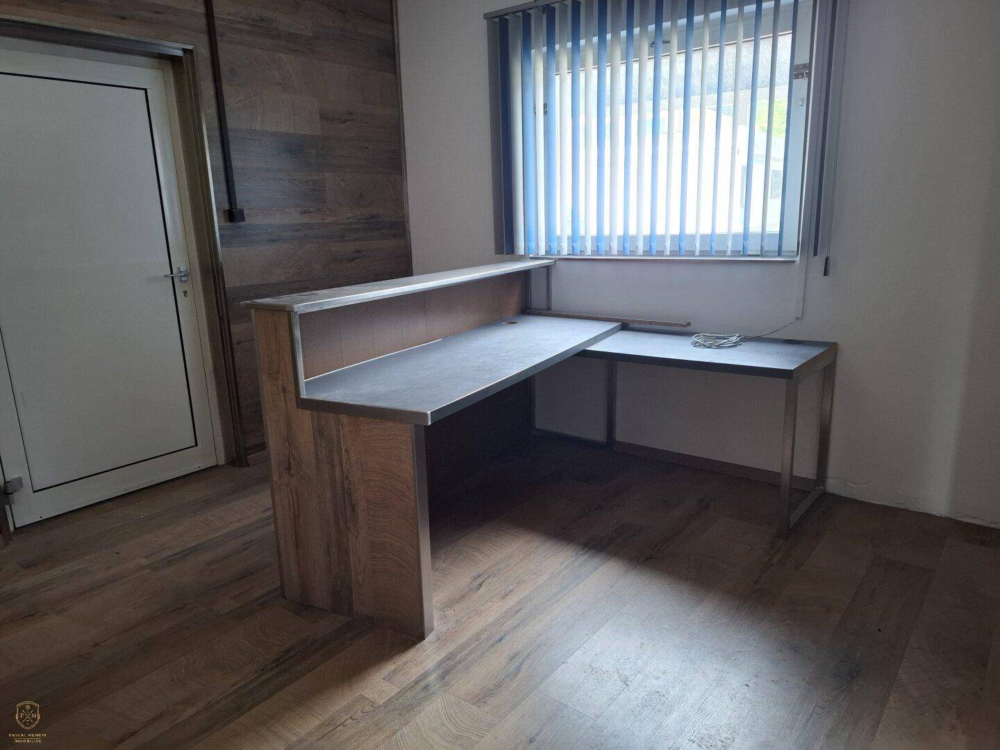 Empfangsbereich oder Büro mit Holzboden, Fenster mit Jalousien und einem L-förmigen Schreibtisch.