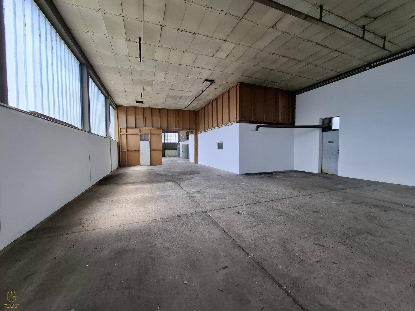 Geräumiger Lagerbereich mit Betonboden, hohen Fenstern und teilweise holzverkleideten Wänden im hinteren Bereich.