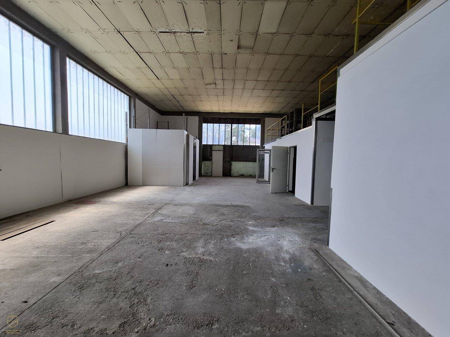 Großer, leerer Industrieraum mit Betonboden, hohen Fenstern und abgetrennten Bereichen im Hintergrund.