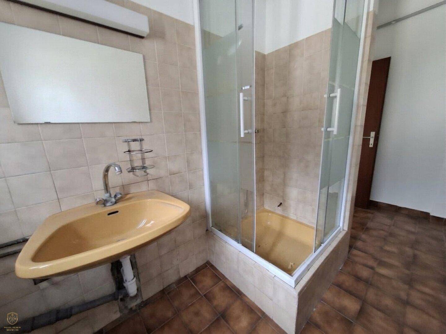 Einfaches Badezimmer mit beigefarbenen Fliesen, einer Duschkabine und einem gelblichen Waschbecken.