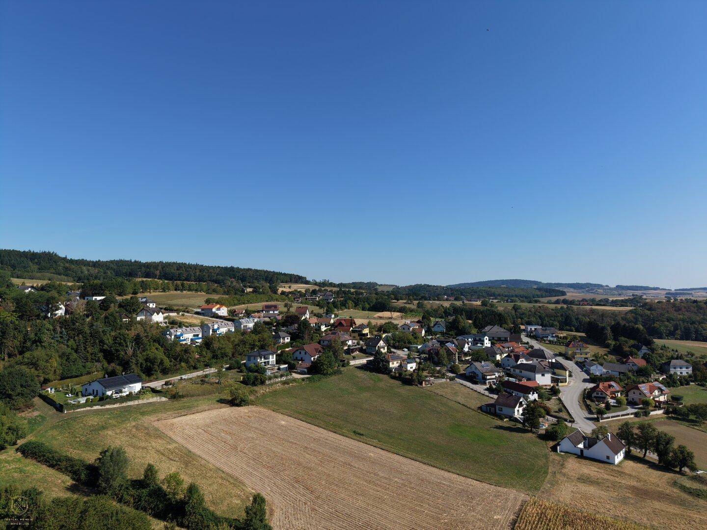 Weitläufige Luftaufnahme einer ländlichen Gegend mit verstreuten Häusern, Feldern und Wäldern unter blauem Himmel.
