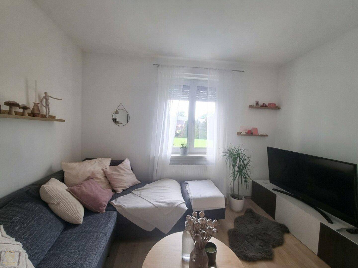 Gemütliches Wohnzimmer mit Sofa, Fernseher und hellem Fenster, das viel Tageslicht hereinlässt.
