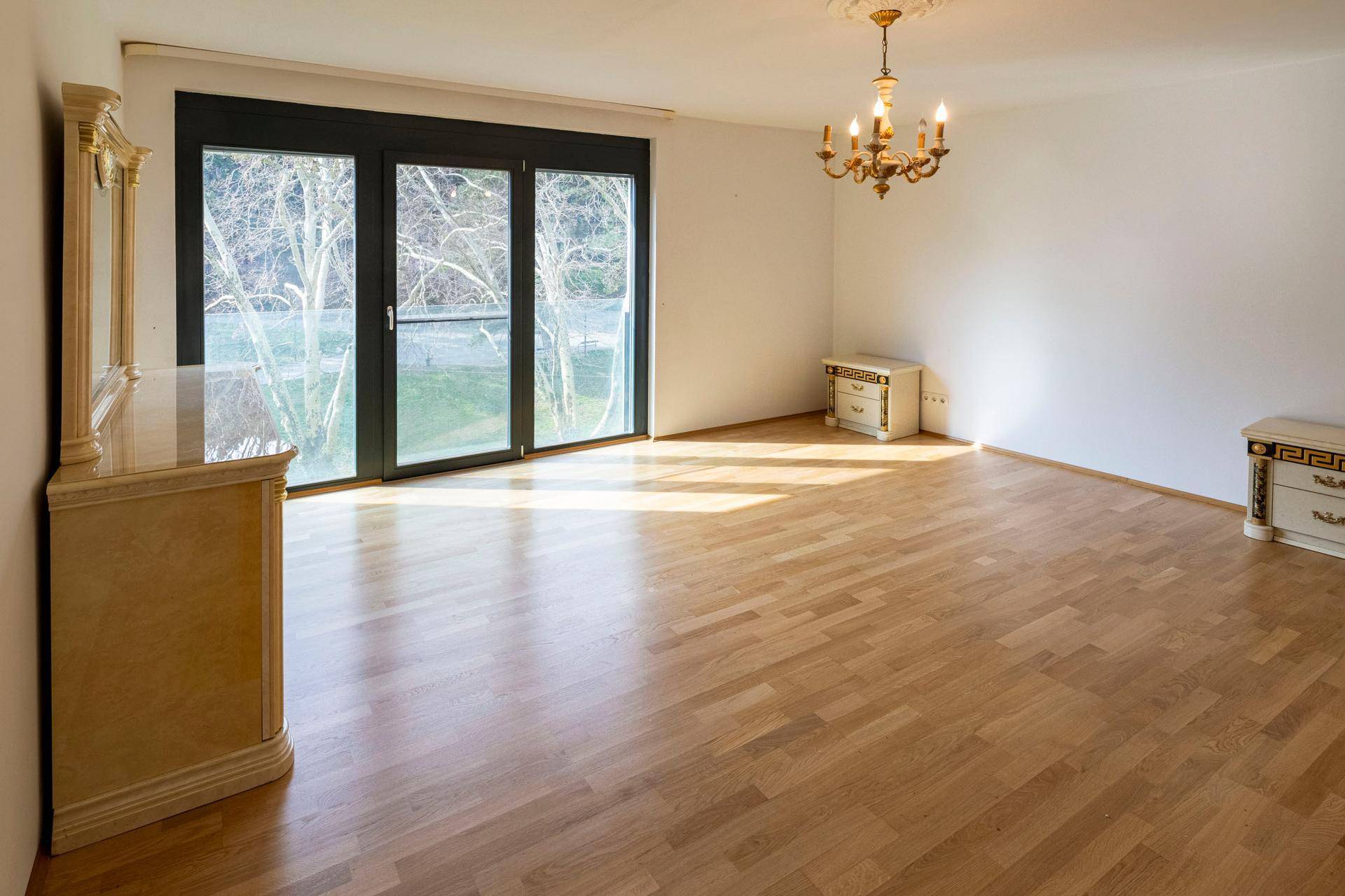 Leeres Schlafzimmer mit Parkettboden, großen Fenstern und klassischem Beistellschrank.