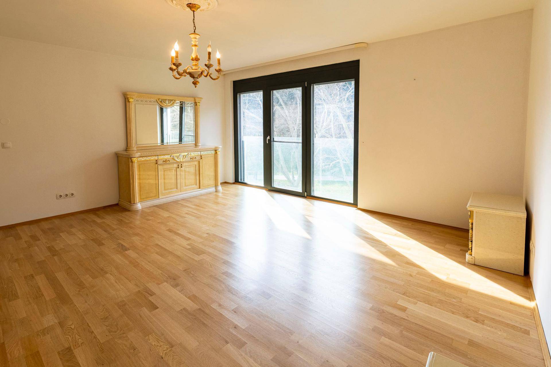 Geräumiger Raum mit Parkettboden, großen Fenstern und klassischem Sideboard.