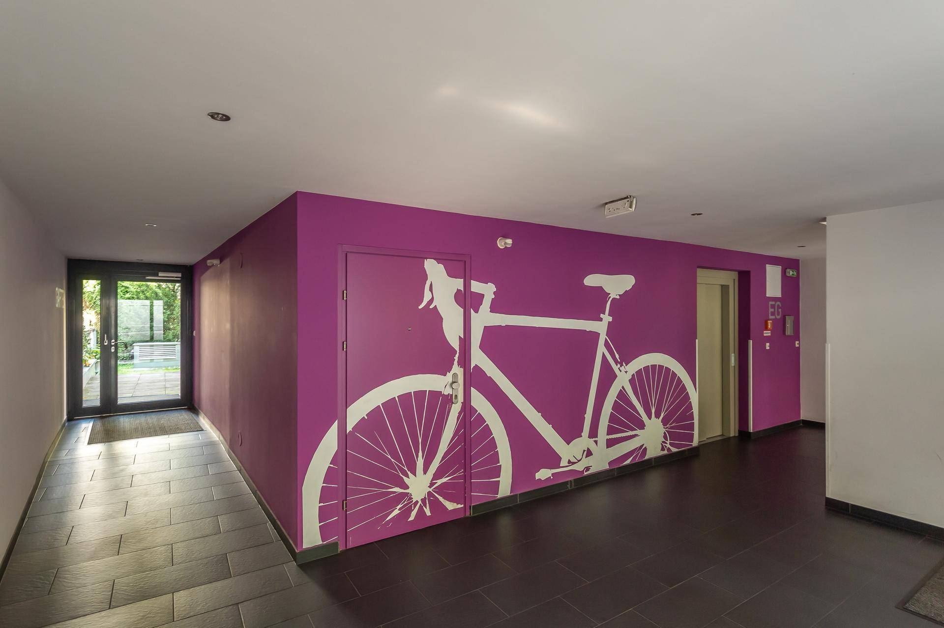 Heller Eingangsbereich mit dunklen Fliesen, lila Wand und Fahrrad-Wandbild.