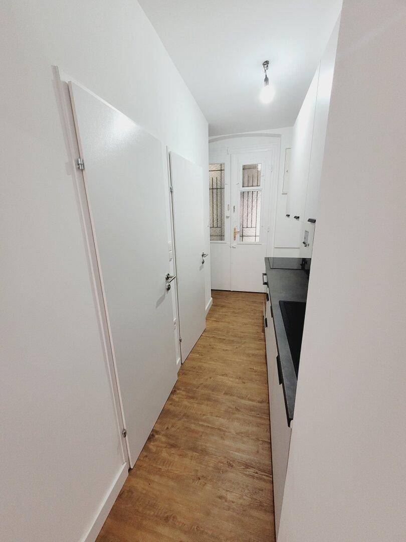 Schmale, moderne Küche mit weißen Schränken, dunkler Arbeitsplatte und Holzboden, führt zu einem Ausgang.