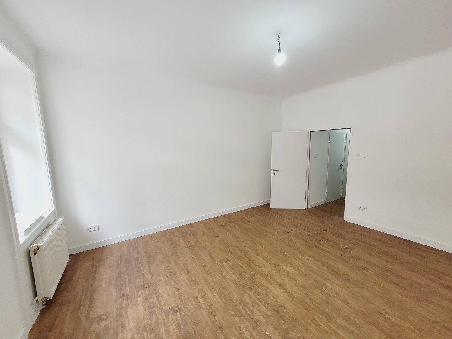 Geräumiges Zimmer mit Holzboden und weiß gestrichenen Wänden, mit Zugang zu weiteren Räumen.
