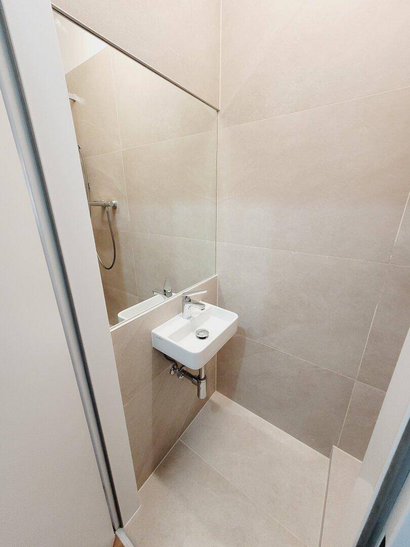 Modernes Badezimmer mit beigefarbenen Fliesen, kleinem Waschbecken und großem Spiegel.